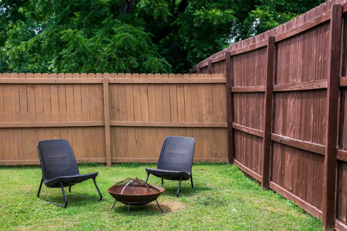 An inviting backyard scene featuring two outdoor chairs, a fire pit, a wooden fence, and a grassy lawn. This private, fully fenced outdoor area offers a safe place for children to play and a cozy spot for families to gather around the fire pit.
