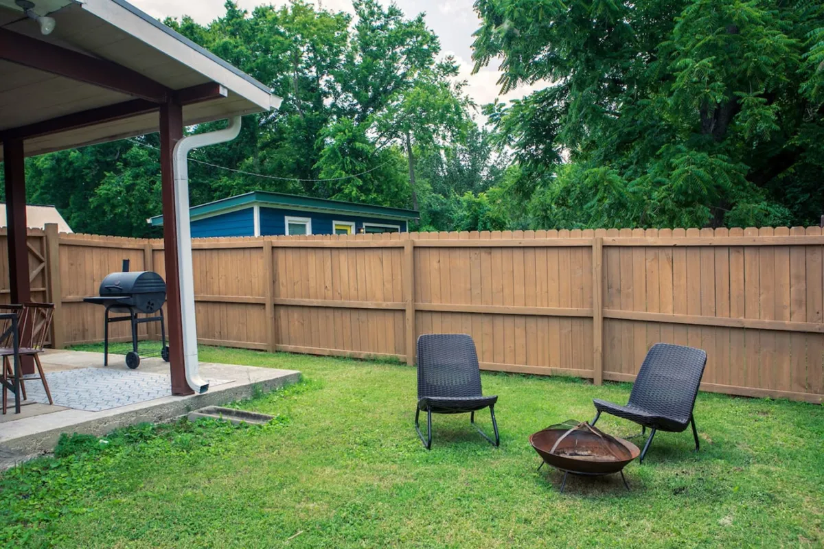 Set in a spacious, fenced yard, this image displays a grill, fire pit, and lawn chairs. The scene exudes a laid-back atmosphere ideal for hosting outdoor gatherings, relaxing by the fire, or simply enjoying the beauty of the fenced backyard.