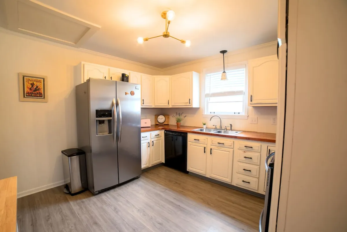 A kitchen view focusing on a stainless steel refrigerator, black dishwasher, wooden countertops, a large kitchen sink, and a hanging light fixture. The setup exudes warmth and practicality, with plenty of space for cooking, gathering ingredients, and sharing moments with family or friends.