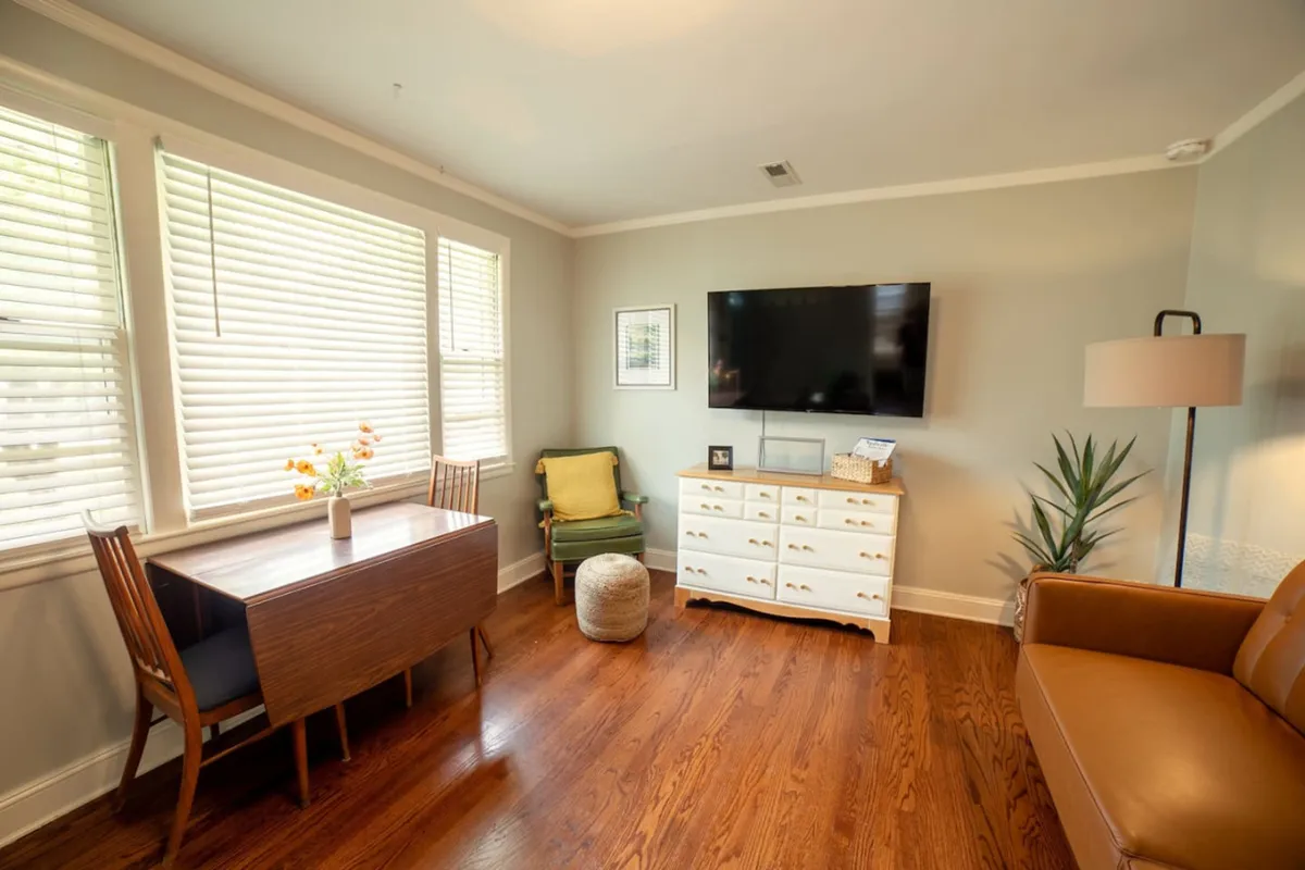 A cozy and inviting living and dining area, featuring a central dining table, a comfortable armchair, a television perfect for movie nights, and a lush plant that adds a touch of greenery. The open-plan layout is visible, blending relaxation and communal dining spaces seamlessly.