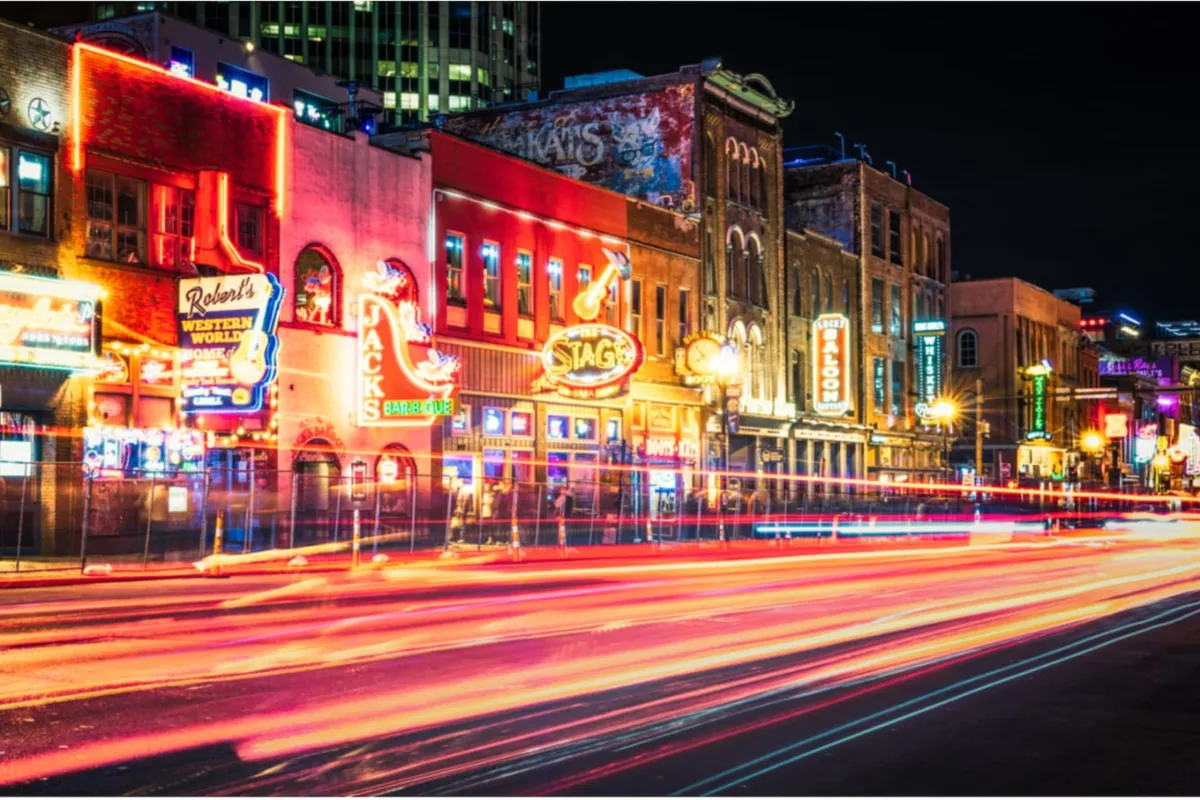 A lively Nashville street scene featuring glowing neon signs, historic buildings, and a bustling crowd enjoying live music, dining, and nightlife. The vibrant energy and iconic city atmosphere are immediately recognizable, capturing the essence of downtown Nashville's entertainment district.