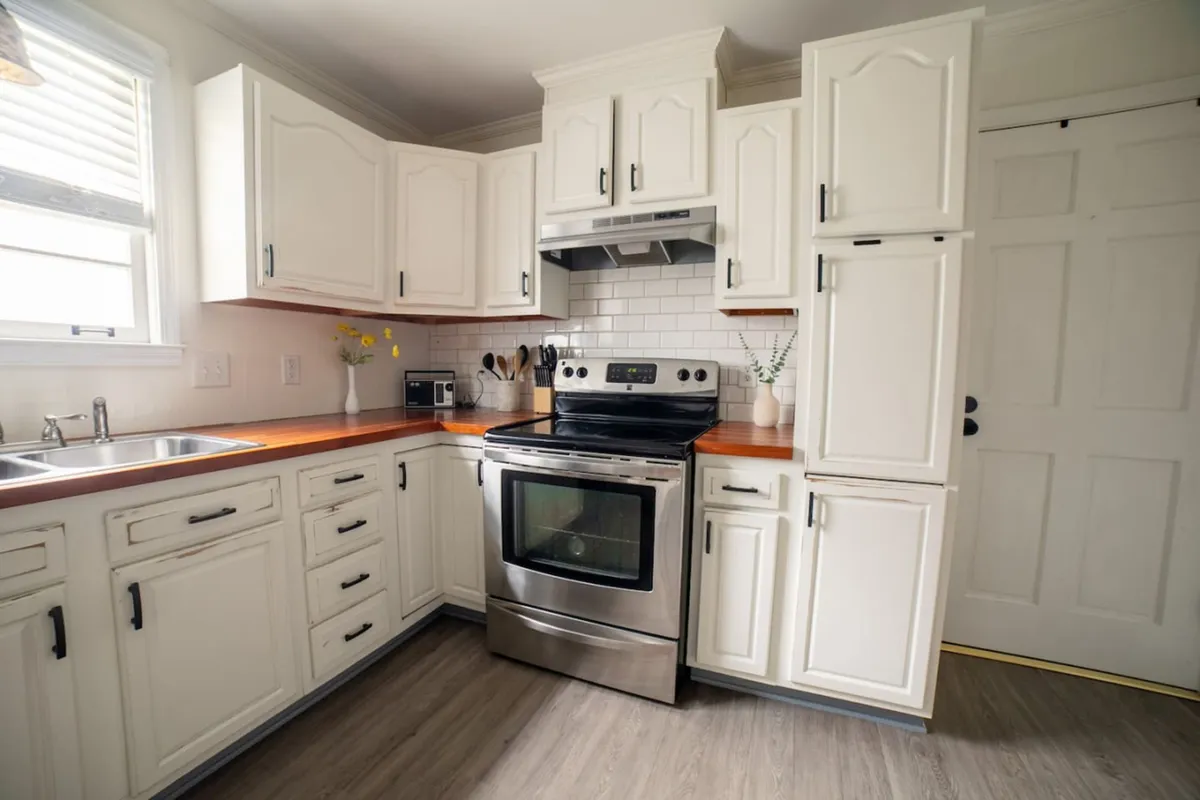 A warm, welcoming kitchen featuring a stainless steel oven, wooden countertops, white cabinetry, a sink beneath a window, and a decorative vase with fresh flowers. The space is bathed in natural light, with ample counter space for meal preparation and a cozy, homey feel ideal for cooking and gathering.