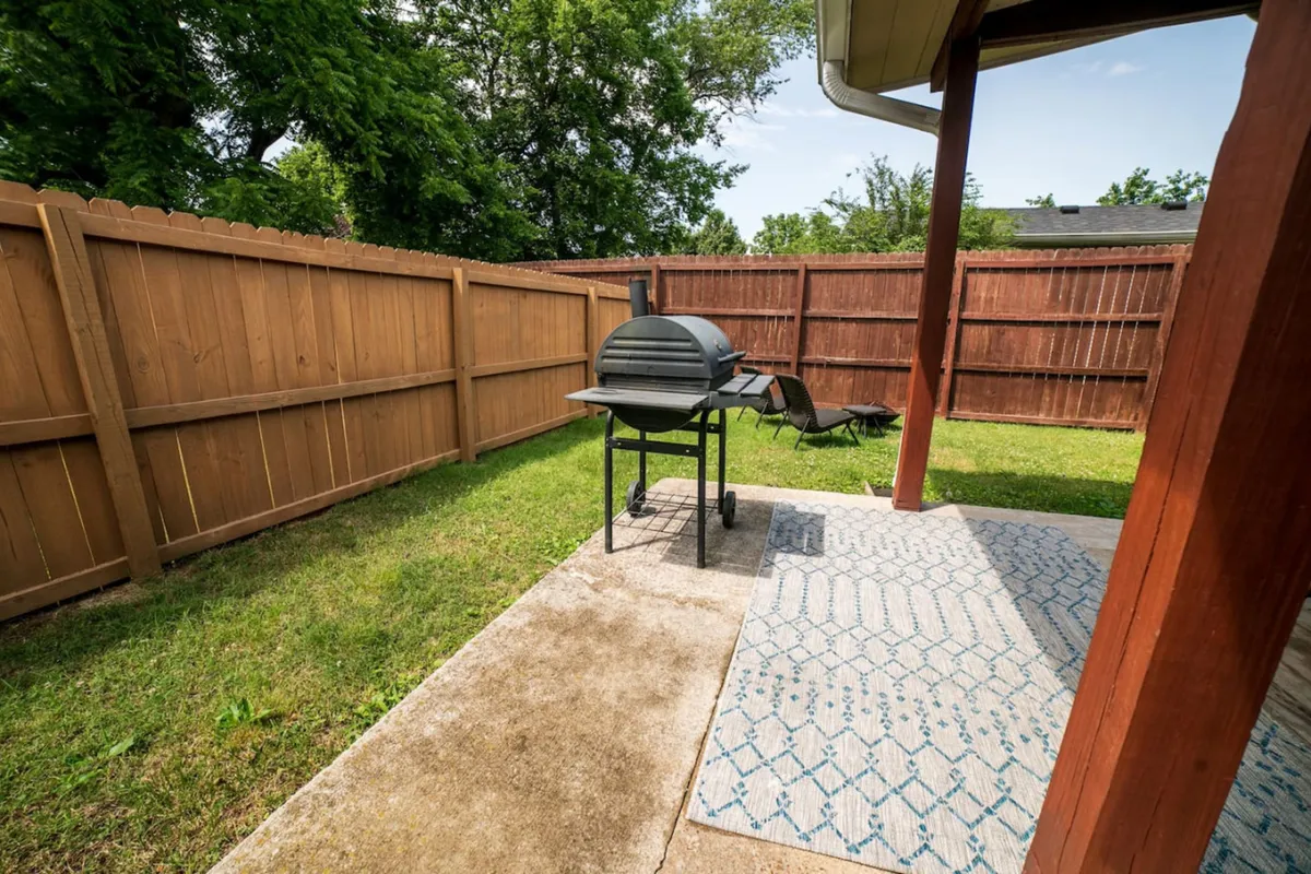 A casual backyard scene showing a grill, colorful outdoor rug, green lawn, and the privacy of a surrounding fence. The setup is perfect for weekend barbecues, lounging, or entertaining friends and family.