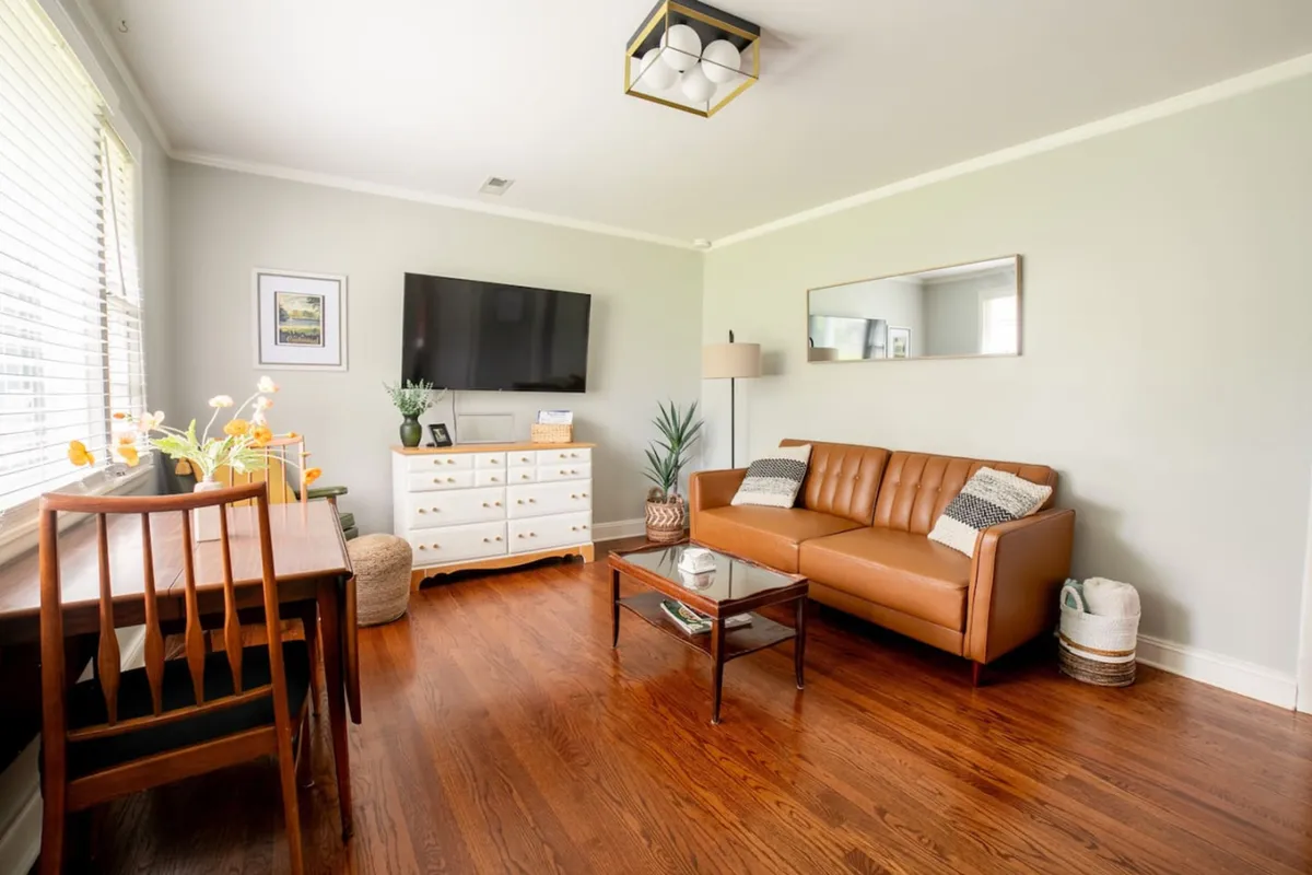 A welcoming living and dining area featuring a plush leather sofa, stylish coffee table, cozy dining table for four, decorative greenery, and a large TV—perfect for relaxing or spending quality time with loved ones.