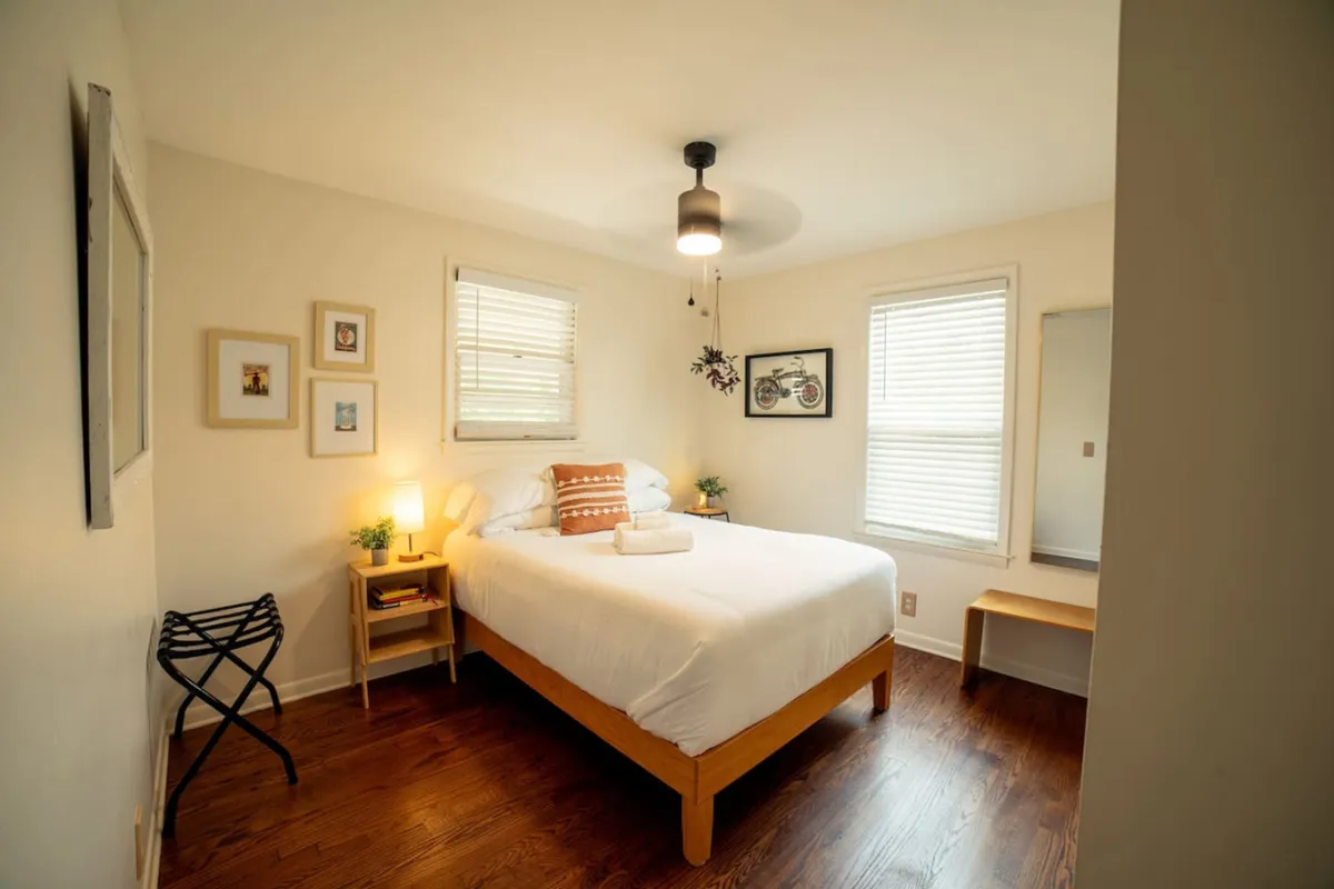 A cozy, sunlit bedroom featuring a comfortable bed adorned with plush bedding. The walls are decorated with tasteful artwork, and decorative plants add a touch of tranquility. A mirror reflects the natural light streaming in, enhancing the room's open and restful atmosphere.