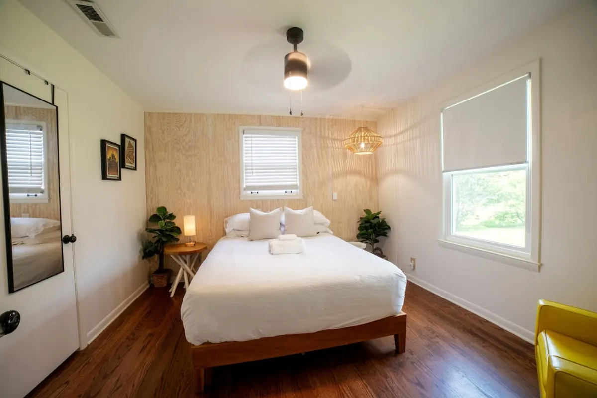A tranquil bedroom offering a serene atmosphere with a welcoming bed, a sunny window, a cheerful yellow chair, and artful wall decor. The warm light creates a soothing space ideal for unwinding, reading, or catching up on rest.